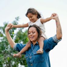 Northside Vistas apartments resident carrying a smiling child on her shoulders outdoors, both holding hands, with greenery in the background.