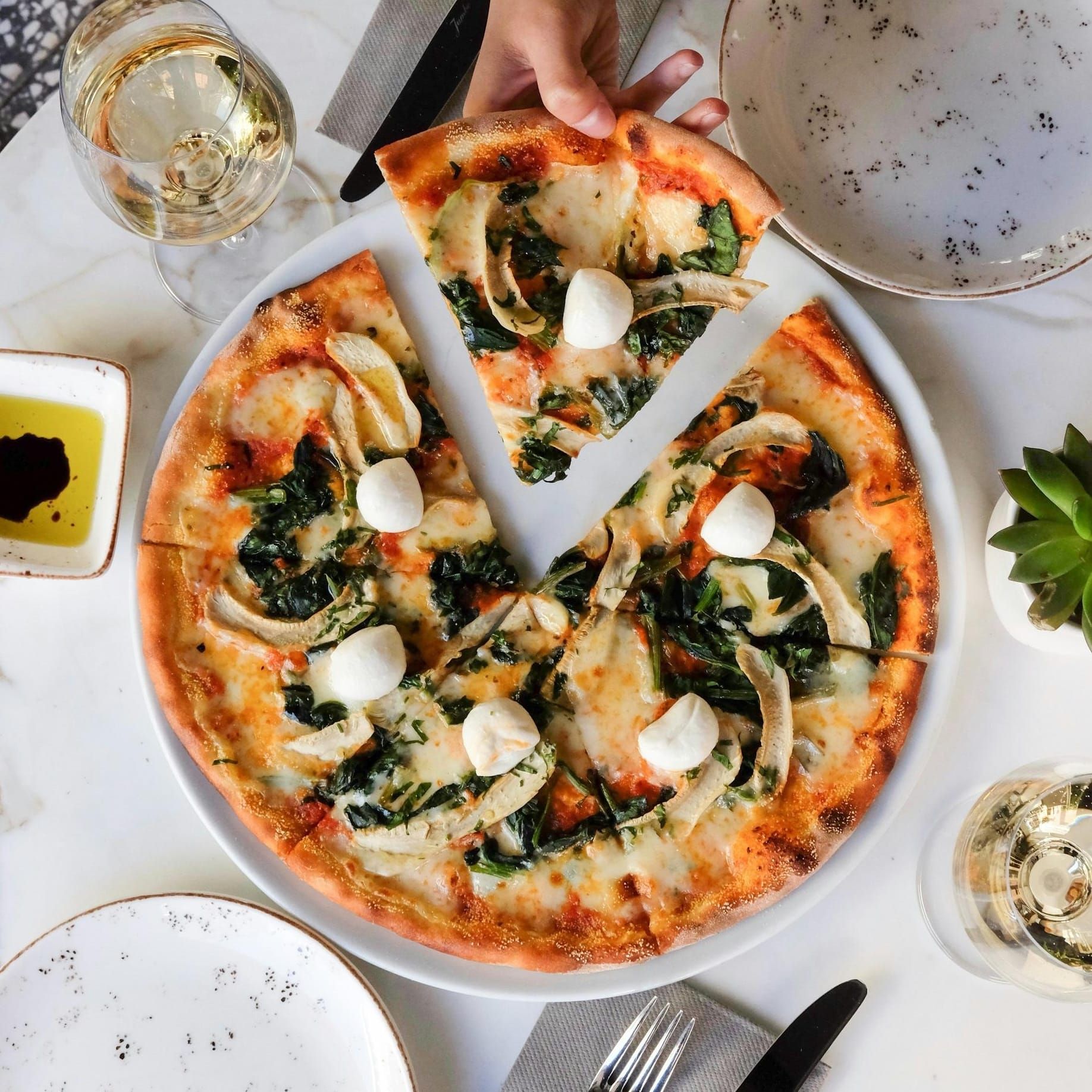 Hand taking a slice of spinach and cheese pizza on a table set with wine near Northside Vistas apartments.
